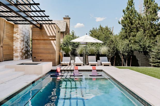 A bright, contemporary backyard pool area with a long rectangular swimming pool, pale stone paving, built-in steps, and a shallow raised water feature. A timber-clad and brick house borders the space, with a black metal pergola casting strong linear shadows. Poolside loungers, an umbrella, screening plants, lawn, and mature trees create a refined outdoor entertaining setting.