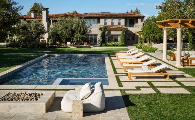 A landscaped residential courtyard with a rectangular swimming pool, paved terrace, white loungers, and a covered outdoor seating area. The two-storey house has a warm stucco facade, red clay tiled roof, arched and divided-light openings, masonry chimneys, and Mediterranean-inspired detailing. Geometric paving and clipped lawns frame the pool and garden.