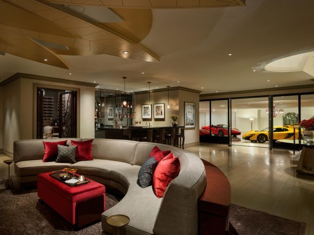A luxurious open-plan living room with a large curved sectional sofa, red upholstered ottoman, dark timber accents, framed artwork, and warm recessed lighting. A glass-enclosed car display garage is visible beyond the living area, containing brightly coloured sports cars. The room features an ornate sculptural ceiling, polished timber flooring, and a refined, dramatic atmosphere.