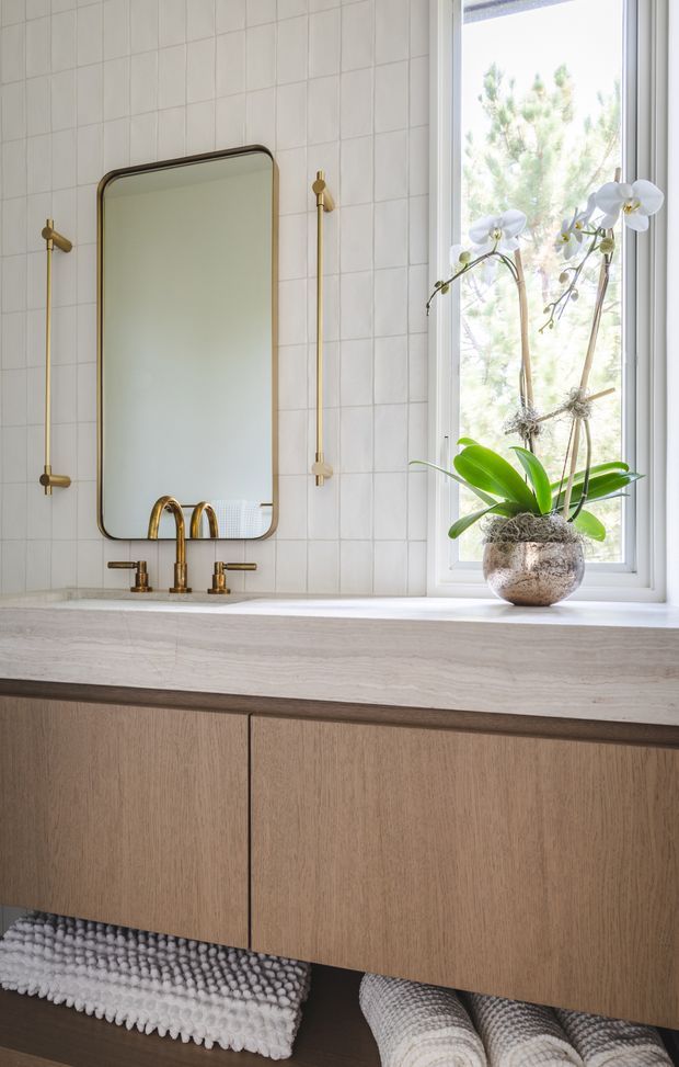 A bright, refined bathroom vanity with a long pale stone benchtop, warm timber floating cabinetry, white square wall tiles, and brushed brass wall-mounted fixtures. A rounded-rectangle mirror hangs above the double-handle basin tap, while a large window brings in natural light and frames an orchid plant on the counter.