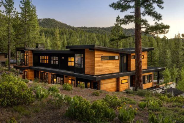 A contemporary two-storey home set among pine and woodland vegetation on a sloping, mountainous site. The building has a low-pitched flat roof, strong horizontal forms, dark charcoal exterior sections, warm timber cladding, large windows, and covered terraces or balconies.