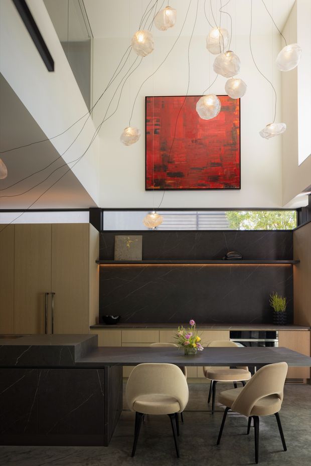A sophisticated contemporary kitchen and dining area with a dramatic double-height ceiling. Dark veined stone forms the island, splashback, and benchtops, contrasted with warm timber cabinetry and pale upholstered dining chairs. A sculptural cluster of suspended glass pendant lights hangs above, while a large red abstract artwork provides a bold focal point. Clerestory-style glazing brings in natural light above the joinery.