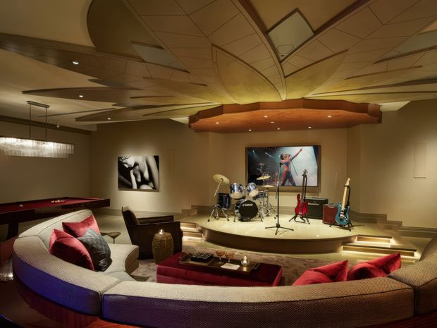 A luxurious, warmly lit home media and music room with a curved sectional sofa surrounding a low coffee table, a raised stage holding a drum kit and several guitars, a wall-mounted screen, a pool table at left, and distinctive curved timber ceiling detailing with recessed lighting.