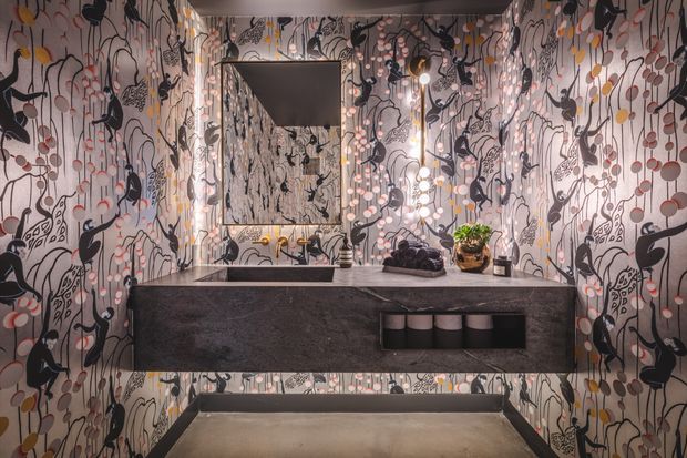 A dramatic, compact bathroom vanity area enclosed in floor-to-ceiling illustrated wallpaper featuring abstract botanical and figurative motifs in black, grey, white, blush, and mustard tones. A dark stone floating vanity with integrated basin and wall-mounted tap sits beneath a rectangular mirror, with warm decorative lighting and small plants adding detail. The space has a polished, eclectic and highly expressive interior aesthetic.