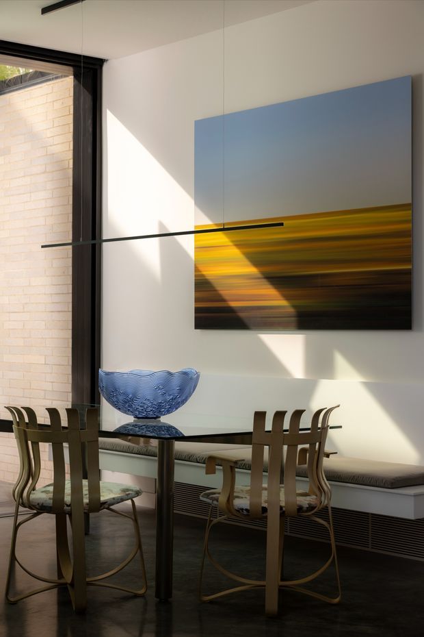 A sunlit dining area with a round glass-topped dining table, sculptural wood-toned chairs, and a built-in window bench. A large abstract artwork in blue, yellow, and dark tones hangs on the white wall, while a blue glass bowl forms a tabletop centerpiece. Full-height black-framed glazing opens to a pale brick exterior wall, and linear black pendant lighting crosses the room. The space has a refined, art-focused contemporary character with strong natural light and dark polished flooring.