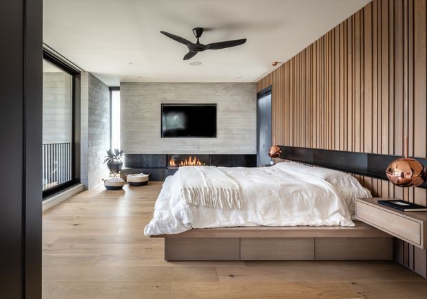 A contemporary master bedroom with a low platform bed and integrated timber storage, facing a textured light stone feature wall with a wall-mounted television and linear fireplace. Vertical timber wall battens form a warm feature wall behind the bed, with black detailing, copper-toned bedside lamps, pale timber flooring, floor-to-ceiling glazing, and a ceiling fan.