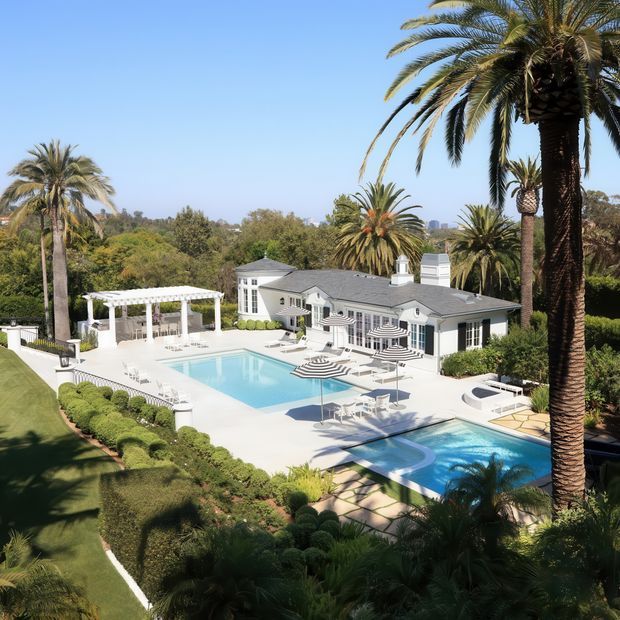Aerial view of a landscaped residential property with a large white-painted, low-rise house, extensive white pool terrace, two swimming pools, striped umbrellas, manicured hedges, a white pergola, and tall palm trees. The home has a restrained traditional character with a dark low-pitched roof, multi-pane windows, shutters, and covered outdoor areas. The scene is bright, warm, and resort-like.