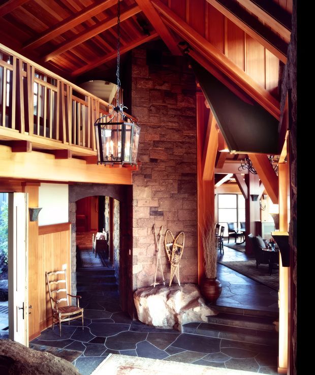Rustic lodge-style interior entry with a dramatic exposed timber roof structure, mezzanine balcony, central rough-stone fireplace or chimney mass, dark irregular stone flooring, and warm timber joinery. A lantern pendant hangs over the entry, with views into adjoining living and dining areas.