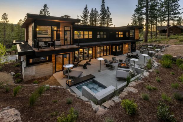 A large contemporary two-storey dark-clad house set into a wooded, sloping site. The home features extensive floor-to-ceiling glazing, projecting balconies and terraces, warm interior lighting, and a landscaped outdoor entertaining area with multiple lounge and dining settings, a fire feature, and a small linear pool or spa. Tall conifer trees surround the property.