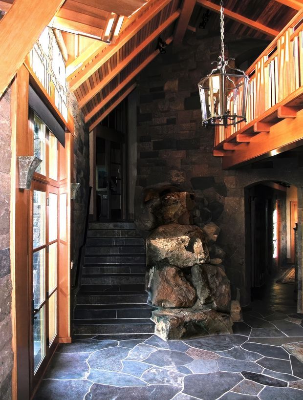 A dramatic double-height interior entry hall with a rustic stone feature wall and integrated natural rock formation, dark slate flagstone flooring, timber-framed glazing, exposed timber roof structure, and an upper-level timber balcony. A large hanging lantern illuminates the lodge-like space, with stairs rising along the left side.