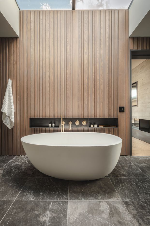 A refined contemporary bathroom featuring a large freestanding white oval bathtub centered against a full-height vertical timber slat wall. A dark built-in recessed shelf with brass-toned tapware and bathroom accessories runs behind the bath. The room has dark grey stone-look floor tiles, a roof skylight above, and an opening to an adjacent space at right.