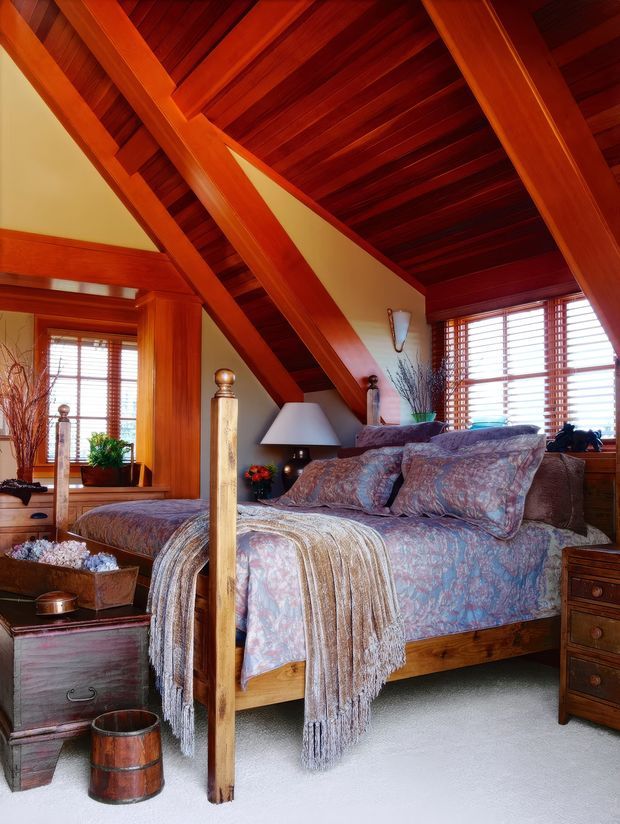 A richly detailed bedroom beneath a dramatic timber-framed sloping ceiling. The room features exposed dark timber roof boards and substantial polished timber beams, warm wood-framed windows with horizontal blinds, a four-poster bed dressed in a muted floral coverlet and layered throw, bedside furniture, lamps, and natural decorative branches. The palette is warm and earthy with strong red-brown timber contrasted against pale walls and carpet.