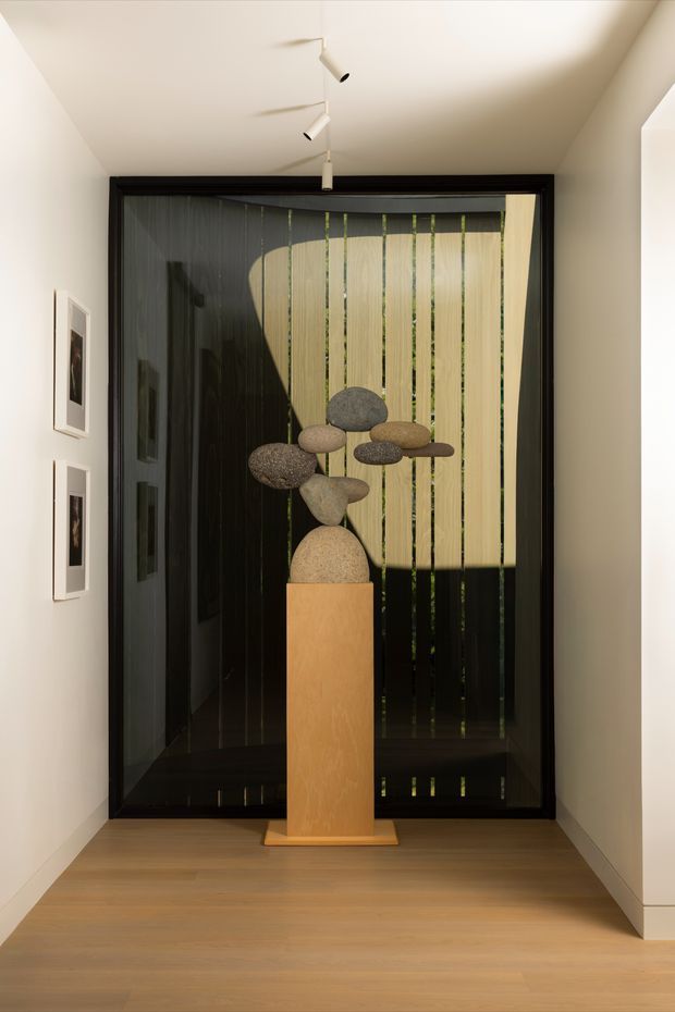 A refined residential hallway or entryway with pale timber flooring, white walls, and a large black-framed glazed opening. A sculptural arrangement of smooth natural stones sits on a warm timber pedestal in front of the glazing, with vertical timber screening and dark reflective surfaces beyond. Minimal track lighting and framed artwork reinforce the restrained, gallery-like atmosphere.