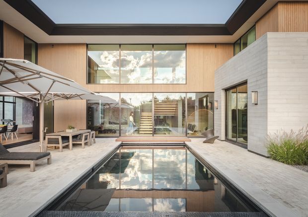 A contemporary two-storey courtyard house arranged around a narrow reflecting pool. The open courtyard features pale paved terraces, a dining and lounge area, large glazed openings, warm vertical timber wall cladding, dark roof framing, and a prominent pale masonry volume. A central staircase and extensive glazing connect the courtyard to the interior, creating strong reflections in the pool.