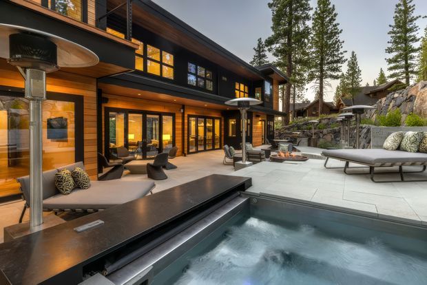A sophisticated multi-level mountain home opens onto a generous paved terrace and spa pool. The dark exterior cladding contrasts with warm timber soffits, extensive glazing, and glowing interior lighting. Contemporary outdoor seating, patio heaters, a long linear water feature or spa edge, and stepped landscaping with exposed rock create an inviting resort-like outdoor entertaining space.