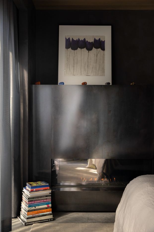 A dark, moody bedroom interior with a partially visible bed in the foreground, a dramatic black or charcoal feature wall, and a large reflective built-in fireplace or firebox integrated into dark cabinetry. A stack of design books sits beside the fireplace, while sheer curtains filter light at the window. Framed botanical artwork and small decorative objects rest above the fireplace enclosure. The room has a refined, monochromatic scheme with warm firelight and subtle metallic reflections.