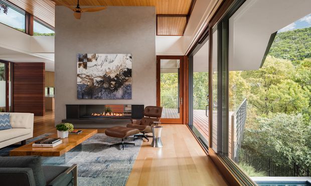 A refined contemporary living room with a dramatic double-height or vaulted volume, expansive floor-to-ceiling glazing, and a timber-lined ceiling. The room features a linear fireplace set into a dark wall, large abstract artwork, a low timber coffee table, neutral sofas, and a leather lounge chair. Glazing opens toward a timber deck and leafy hillside landscape, creating a strong indoor–outdoor connection.