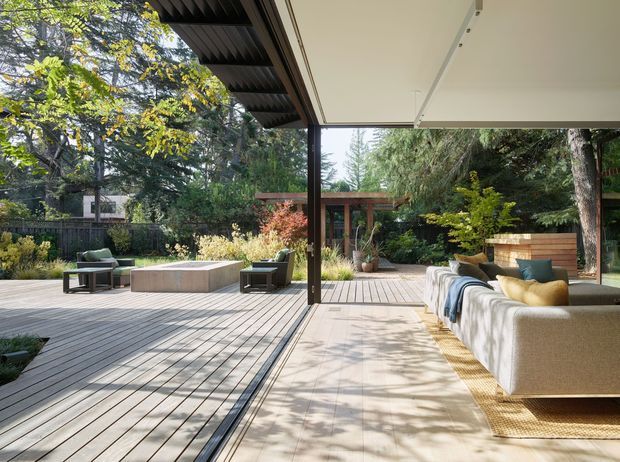A spacious contemporary indoor-outdoor living area opening onto a landscaped garden courtyard. The covered space features a large low-profile upholstered sectional sofa, outdoor lounge chairs, a concrete fire or coffee table, timber decking, light paving, dark slender structural framing, and a broad retractable or glazed opening connecting the interior and exterior. Mature trees and layered planting create a private, natural setting.
