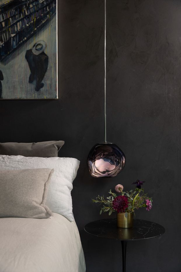 A moody, sophisticated bedroom vignette with a partially visible bed dressed in cream and white linen, set against a dark charcoal textured wall. A reflective smoked-glass pendant hangs beside the bed above a slim black round side table, which holds a brass vase of flowers. A framed artwork is partially visible on the wall.