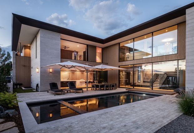 Contemporary two-storey courtyard residence arranged around a rectangular reflecting pool. The home features a dramatic low-pitched roof, white masonry walls, warm vertical timber cladding, extensive floor-to-ceiling glazing, an upper-level balcony, and a paved pool terrace with outdoor seating and umbrellas. The dusk lighting creates a refined, warm atmosphere.