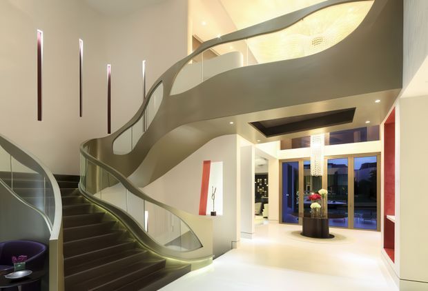 A dramatic, high-end residential entry foyer dominated by a sweeping sculptural staircase with dark treads, curved metallic balustrades, and glass panels. The double-height space has pale walls, recessed lighting, tall narrow feature lights, a contemporary pendant, and expansive blue-framed glazing and doors opening to the exterior. A small seating area and console styling are visible near the stair.
