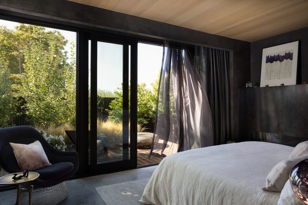 A dark, refined bedroom with a large black-framed glazed sliding door opening to a leafy outdoor area. The room features a low-profile upholstered lounge chair, a neatly made bed with pale bedding, charcoal walls, a warm timber-lined ceiling, sheer and dark curtains, and restrained wall art. Natural light creates a calm contrast between the moody interior and bright garden beyond.