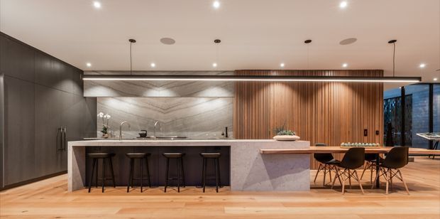 A contemporary open-plan kitchen and dining area with a large stone island, integrated dark cabinetry, timber flooring, a marble-look splashback, and a vertical timber feature wall. The island includes seating for four, while a dining table and chairs sit adjacent beneath warm pendant lighting. Floor-to-ceiling glazing is visible at the right.