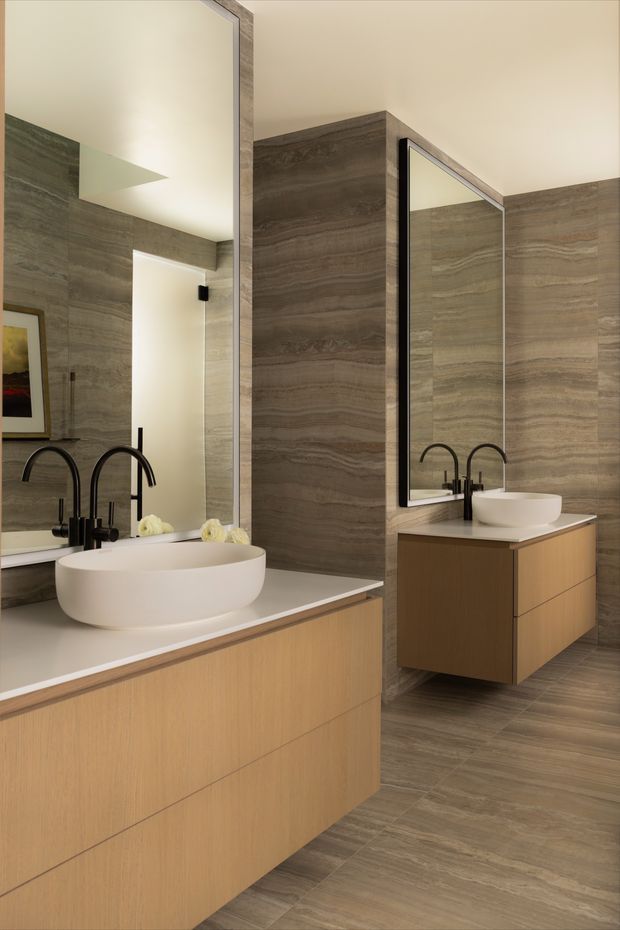 A refined contemporary bathroom featuring a large floating timber-toned vanity with integrated drawers, white vessel basins, matte black curved tapware, and oversized frameless mirrors with dark trim. Grey-brown veined stone-look wall and floor finishes create a cohesive, luxurious material palette, while concealed lighting provides a warm, calm atmosphere.