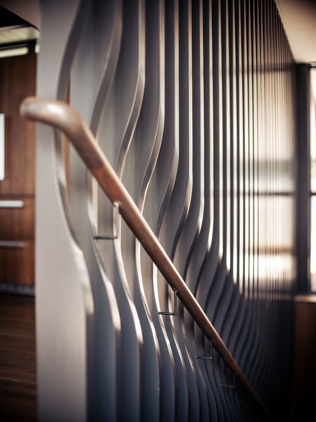 Close-up architectural interior view of a stairway with a curved timber handrail and sculptural, vertically undulating metal balustrade. A dark timber-lined interior and tall linear screened glazing or slatted wall create strong vertical rhythm, with warm natural light and dramatic shadows.