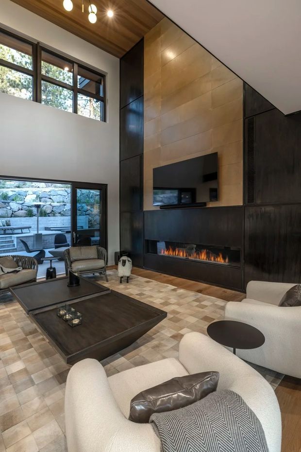 A sophisticated double-height living room centered on a large contemporary fireplace wall. The room features dark built-in wall panelling and cabinetry, a warm timber ceiling and fireplace surround, a wall-mounted television, pale boucle lounge seating, dark low coffee tables, and a patterned tiled floor. Large windows and glazed doors connect the living area to a stone retaining wall and outdoor seating area.