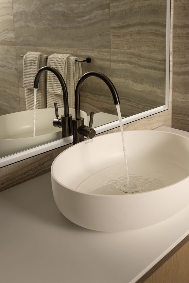 Close-up of a refined bathroom vanity featuring a large white vessel basin on a pale stone or solid-surface benchtop, with a dark brushed-metal mixer tap running water. The wall is clad in richly veined grey-brown stone-look slab tiles, and a large mirror reflects the basin and a towel rail with textured neutral towels. The composition has warm, understated luxury and strong horizontal and organic material textures.