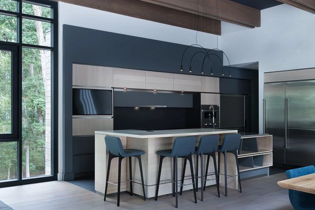 A sophisticated open-plan kitchen with a large island and four blue upholstered bar stools. The kitchen features dark charcoal walls and cabinetry, pale wood-grain overhead and island cabinetry, integrated appliances, a black splashback, and sculptural black pendant lights. Floor-to-ceiling black-framed glazing brings in views of trees, while exposed timber ceiling beams add warmth. Part of a living area with a blue sofa and timber table is visible at the right edge.