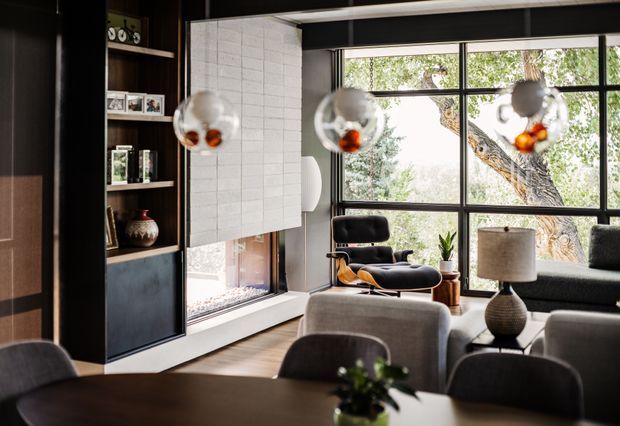 A sophisticated contemporary living room with a dark built-in shelving and cabinetry wall, pale tiled fireplace surround, expansive black-framed windows, and a prominent mid-century lounge chair. A sofa, armchairs, coffee table, dining table and pendant lights create a connected living and dining setting. The room has warm timber flooring, dark joinery, neutral upholstery, and abundant natural light with a leafy tree visible outside.