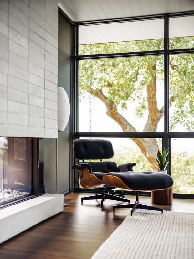 A refined living room corner with a mid-century lounge chair and matching ottoman positioned beside expansive black-framed windows. The room features dark timber flooring, a pale textured masonry fireplace wall with a linear firebox, a neutral textured rug, and a sculptural wall light. Abundant natural light and views of a mature tree create a warm, calm atmosphere.