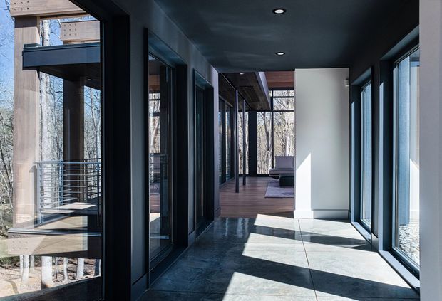 A modern, narrow interior passage or gallery with extensive floor-to-ceiling glazing on both sides, dark ceiling and window frames, pale walls, and large-format grey flooring. The corridor leads toward a timber-floored living area with a purple lounge chair visible in the distance. Exterior woodland and a deck/balustrade are visible through the glazing. Strong daylight creates geometric shadows across the floor.