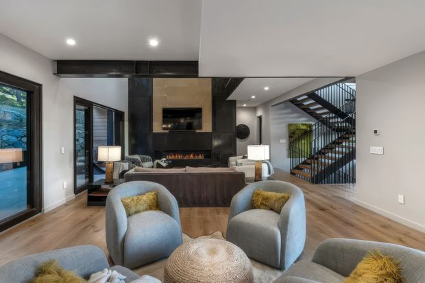 Contemporary open-plan living room with light timber flooring, pale grey walls, and a sophisticated black feature fireplace wall incorporating a television and shelving. The room is furnished with a large brown sofa, pale blue-grey armchairs, patterned cushions, lamps, and a round woven ottoman. Floor-to-ceiling glazing opens toward an outdoor area, while a black metal staircase with timber treads is visible at the rear, creating a strong architectural feature.