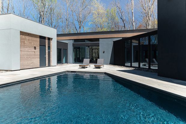 A contemporary single-storey courtyard residence surrounds a rectangular swimming pool. The building has flat rooflines, crisp dark and light rendered walls, extensive black-framed glazing, and a large timber-clad garage or entry door. Pale stone paving frames the pool and connects the wings of the house, with a wooded backdrop beyond.