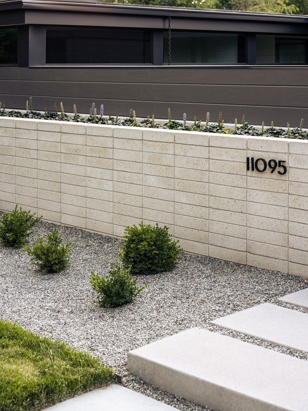 A minimalist residential front boundary with a pale textured concrete block wall bearing the number 11095, dark horizontal-clad upper facade, long dark-framed windows, a narrow planted strip, gravel landscaping with clipped shrubs, and large pale concrete stepping slabs.