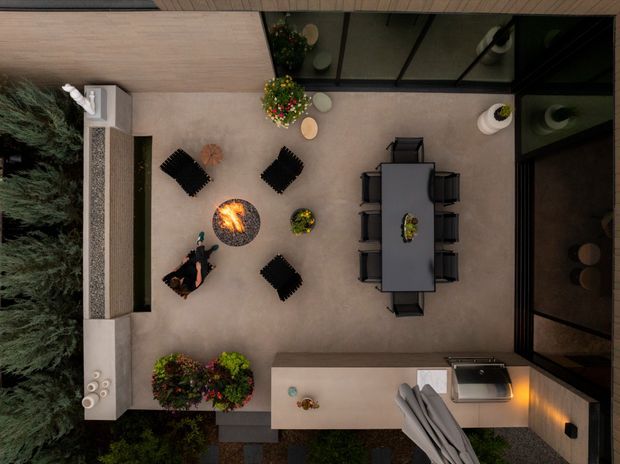 Top-down view of a refined outdoor courtyard entertaining area adjoining a glazed interior. The space includes a long dining table with multiple dark chairs, a circular fire pit with lounge seating, potted plants, built-in perimeter seating or planters, and a covered outdoor kitchen or serving counter. Neutral paving and dark-framed glazing create a contemporary, intimate setting.