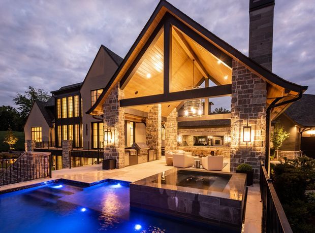 A luxurious multi-level residential home viewed from an outdoor terrace at dusk. The prominent gabled pavilion has a dramatic exposed timber ceiling, heavy dark structural beams, and substantial natural-stone-clad columns and walls. Warm interior and exterior lighting illuminates an outdoor entertaining area with built-in barbecue, seating, a raised square water feature or spa, and a blue-lit swimming pool in the foreground. Dark pitched roofs, large glazed openings, and surrounding landscaped planting contribute to a substantial lodge-like appearance.