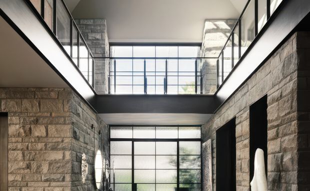 Interior view of a dramatic double-height circulation space with rough-cut stone walls, dark metal-framed glazing, and a glass-balustraded upper-level walkway. Large windows bring in natural light, while black-framed openings and exposed structural geometry create a strong modern architectural character.