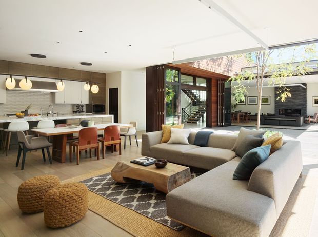 Open-plan contemporary living and dining area with a connected kitchen, featuring a large light upholstered sectional sofa, timber coffee table, patterned rug, dining table with mixed chairs, and a bright white kitchen with island. A dramatic timber-framed glazed opening leads to an additional sitting area and stairway, with abundant natural light and indoor planting.