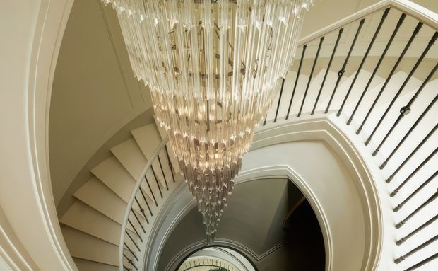 Looking upward through a dramatic curved stairway and landing, with sweeping white architectural arches, dark slender balusters, and a large cascading crystal chandelier suspended through the stairwell void. The image emphasises ornate circulation space, layered curves, and a refined, luxurious atmosphere.