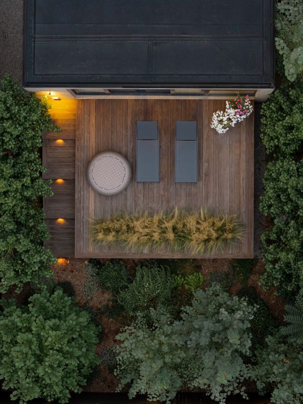 Aerial view of a landscaped outdoor terrace beside a dark-roofed structure. The terrace is clad in warm vertical timber decking and furnished with two slim dark sun loungers and a rounded woven lounge chair. Integrated step lighting glows along the edge, while dense layered planting surrounds the deck and a raised planter of ornamental grasses occupies the foreground.