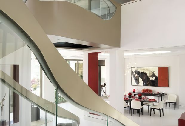 Bright, spacious multi-level contemporary interior featuring a sweeping sculptural staircase with glass balustrades, a curved mezzanine void, extensive glazing, white walls and floors, and a formal dining area with black and white furnishings and red accents. The open-plan circulation space and dining zone create a refined, gallery-like atmosphere.