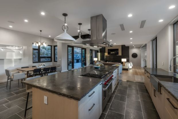 Spacious open-plan kitchen with a large central island, dark stone benchtops, integrated cooktop and oven, light wood cabinetry, pendant lighting, and dark tiled flooring. A dining area and living room are visible beyond the kitchen, with extensive glazing and a contemporary neutral fit-out.