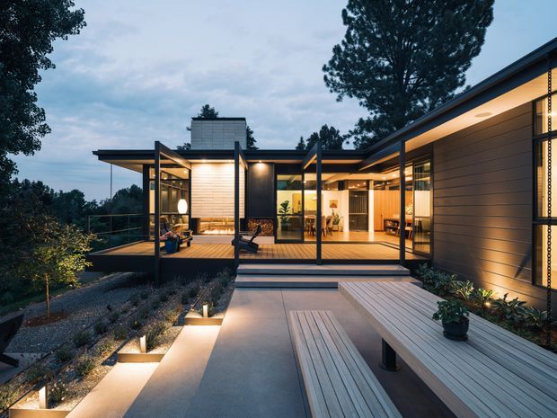 A contemporary single-storey home at dusk, arranged around a broad timber-clad terrace and illuminated outdoor walkway. The building features low-pitched roof planes, extensive floor-to-ceiling glazing, dark slender structural frames, horizontal timber or composite wall cladding, and a warm, open-plan interior visible through the glass. Steps, built-in bench seating, integrated lighting, and landscaped gravel planting connect the house to the surrounding wooded site.