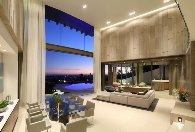 A luxurious double-height living room with floor-to-ceiling glazing overlooking a pool and evening cityscape. The space features a large pale sectional sofa, glass dining table with upholstered chairs, expansive cream porcelain-like flooring, tall sheer curtains, recessed and linear lighting, and a dramatic sculptural timber-panelled feature wall with an upper-level opening and curved glass balustrade. A kitchen or service area is visible beyond the living space.