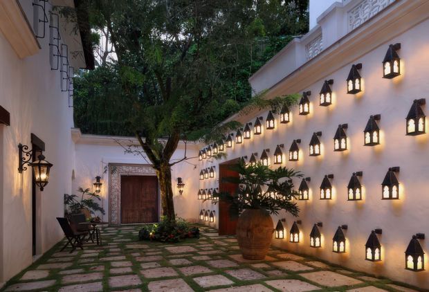 A warmly lit enclosed courtyard with pale rendered walls, decorative parapets, and rows of small lantern-style wall lights. A mature tree, potted tropical planting, patterned stone paving, timber doors, and outdoor seating create an intimate, atmospheric garden retreat.