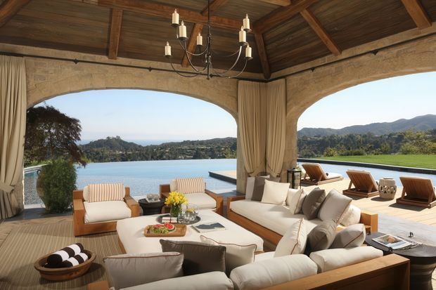 A luxurious open-sided covered outdoor lounge overlooking a swimming pool and expansive lake or water view. The space features a large upholstered sectional sofa, armchairs, coffee table, timber decking, heavy curtains, arched stone openings, and a dark exposed timber-beamed ceiling with a hanging chandelier. Mountainous and landscaped surroundings are visible beyond the terrace.